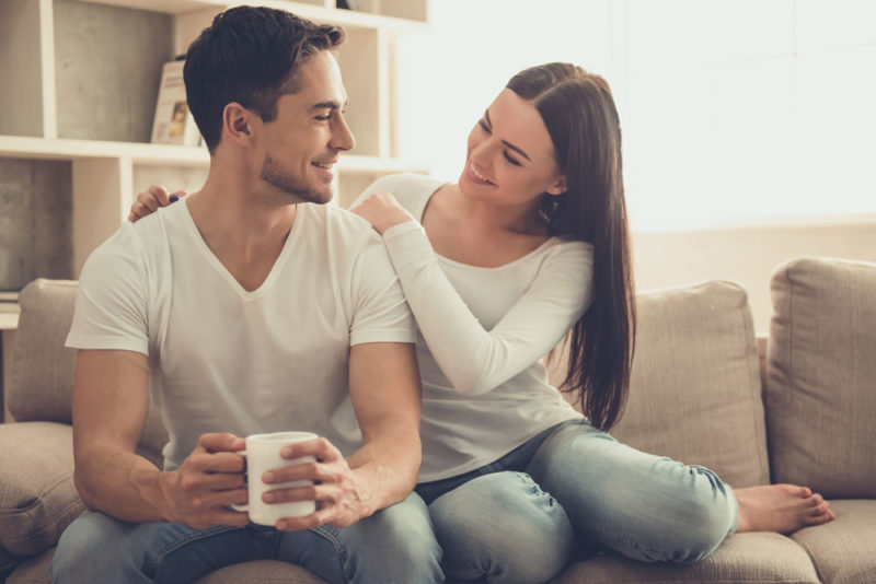 Beautiful young couple is talking and smiling while sitting on sofa at home. Man is holding a cup