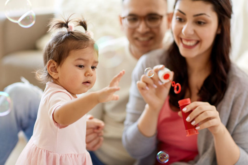 family, childhood and people concept - happy mother blowing soap bubbles and playing with little daughter at home